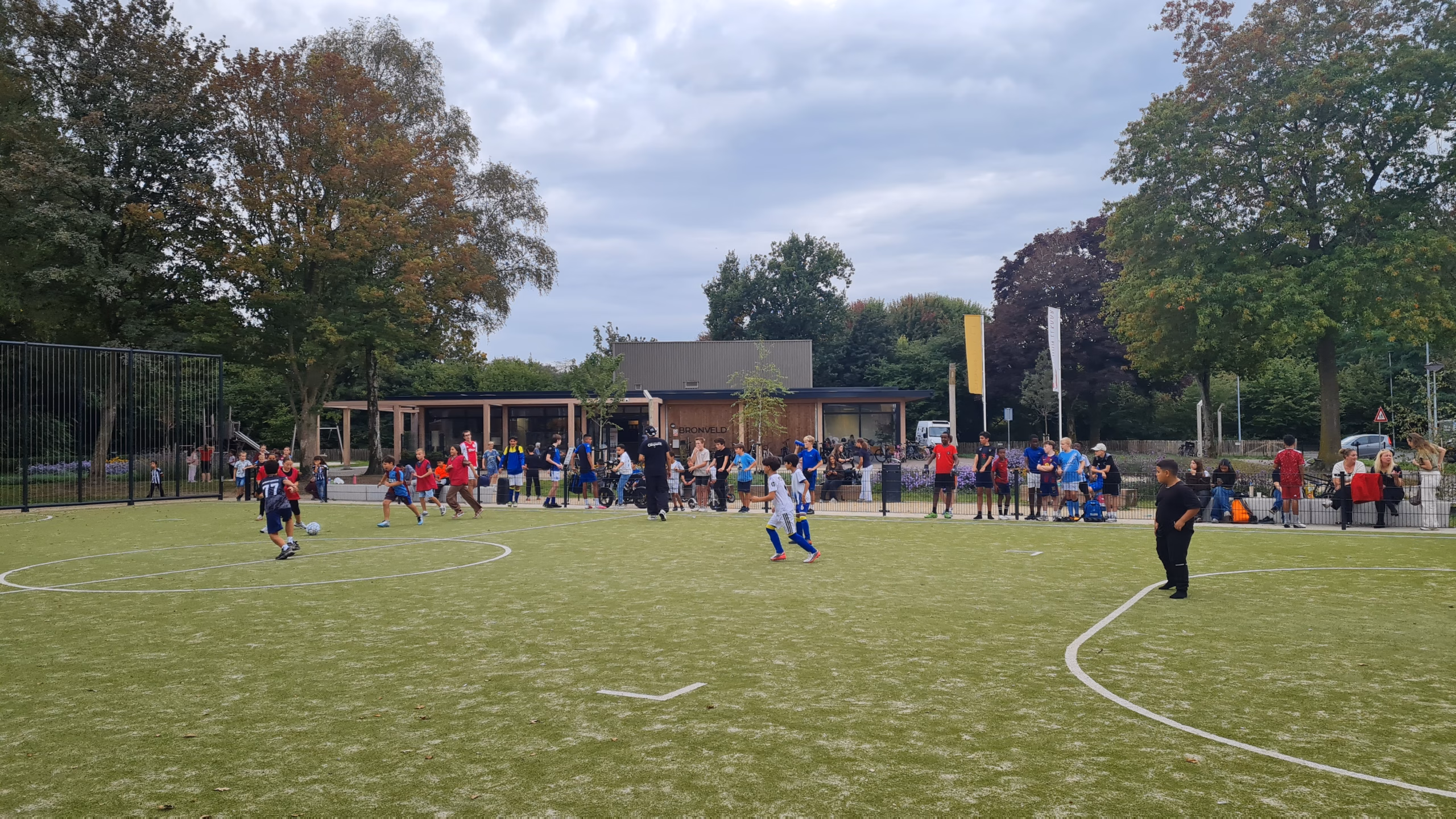 Kinderen voetballen op het veld bij Buurthuis Bronveld, langs het hek kijken buurtbewoners toe.