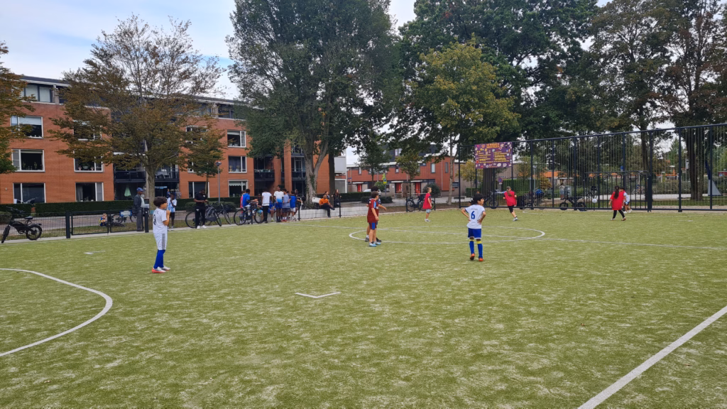 Jonge voetballers in witte en rode shirts op het kunstgrasveld, met woningen aan de overkant op de achtergrond.